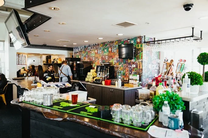 A vibrant bar area featuring a variety of drinks, an artistic wall mural, and patrons enjoying their time.
