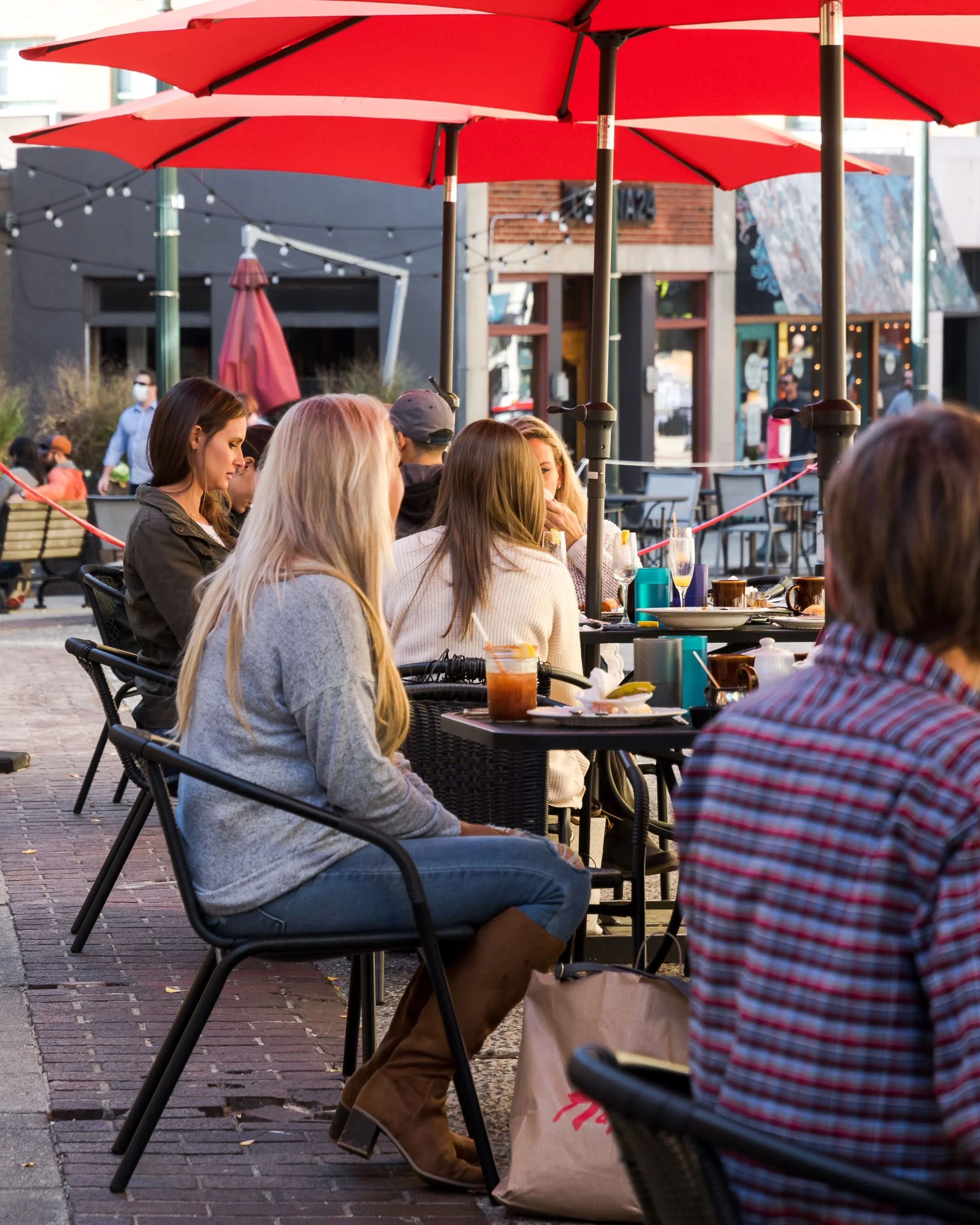 An outdoor dining scene with people seated under red umbrellas, enjoying their meals and drinks in a lively atmosphere.