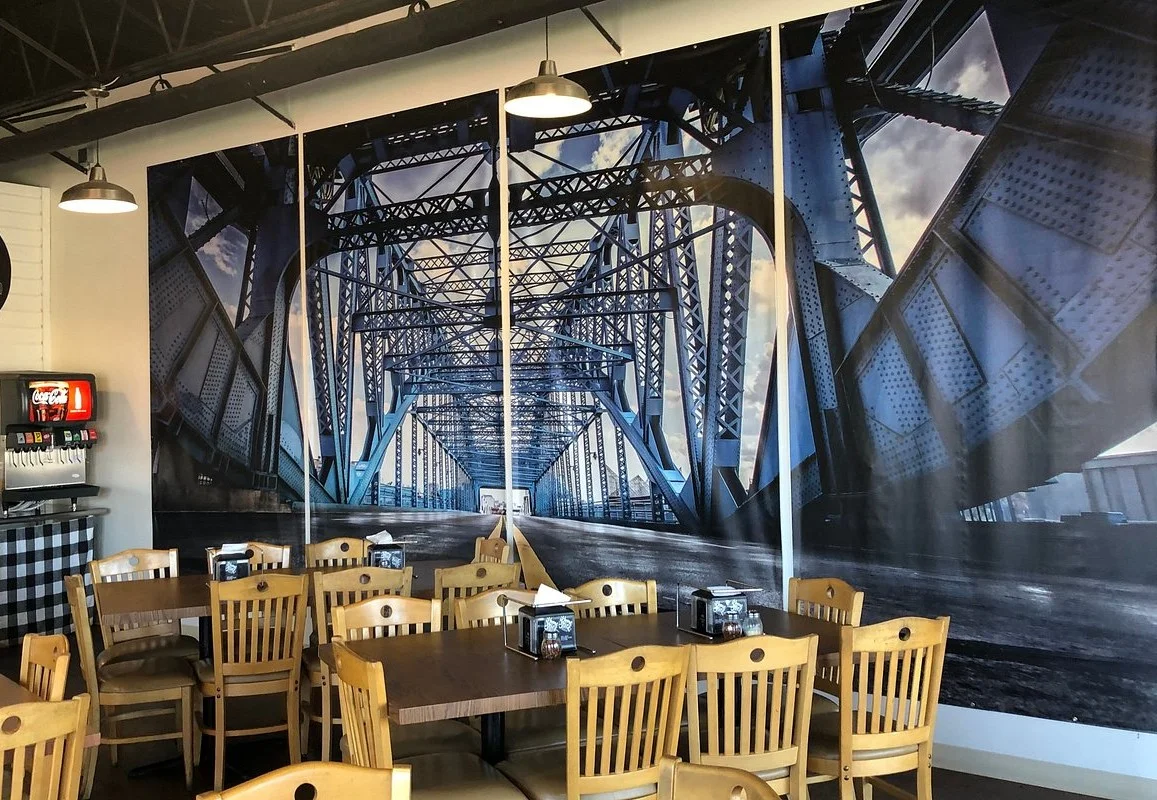 A dining room with wooden chairs and tables, featuring a large mural of a metal bridge on one wall.