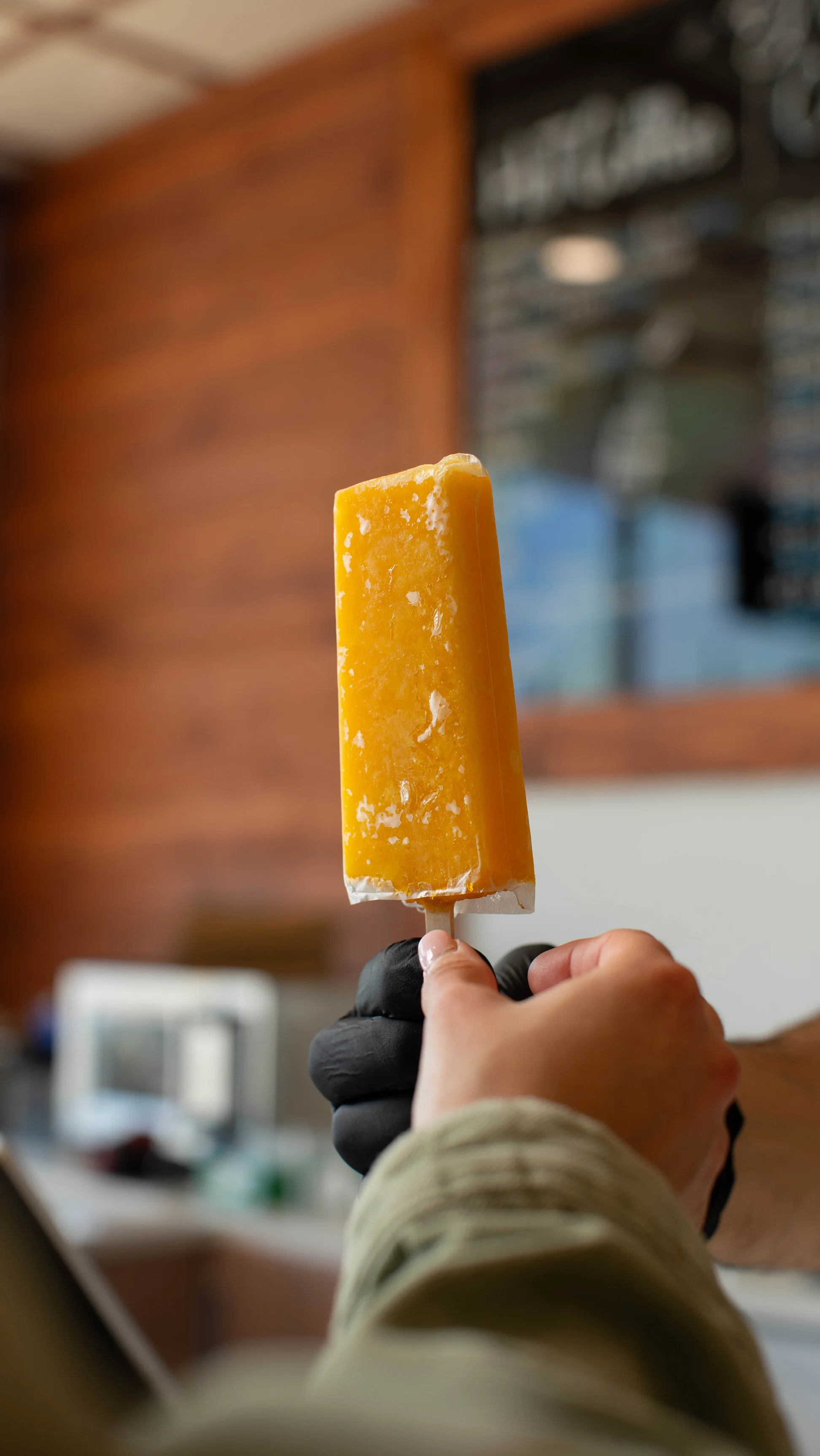 A hand holding a bright yellow popsicle in a café setting.