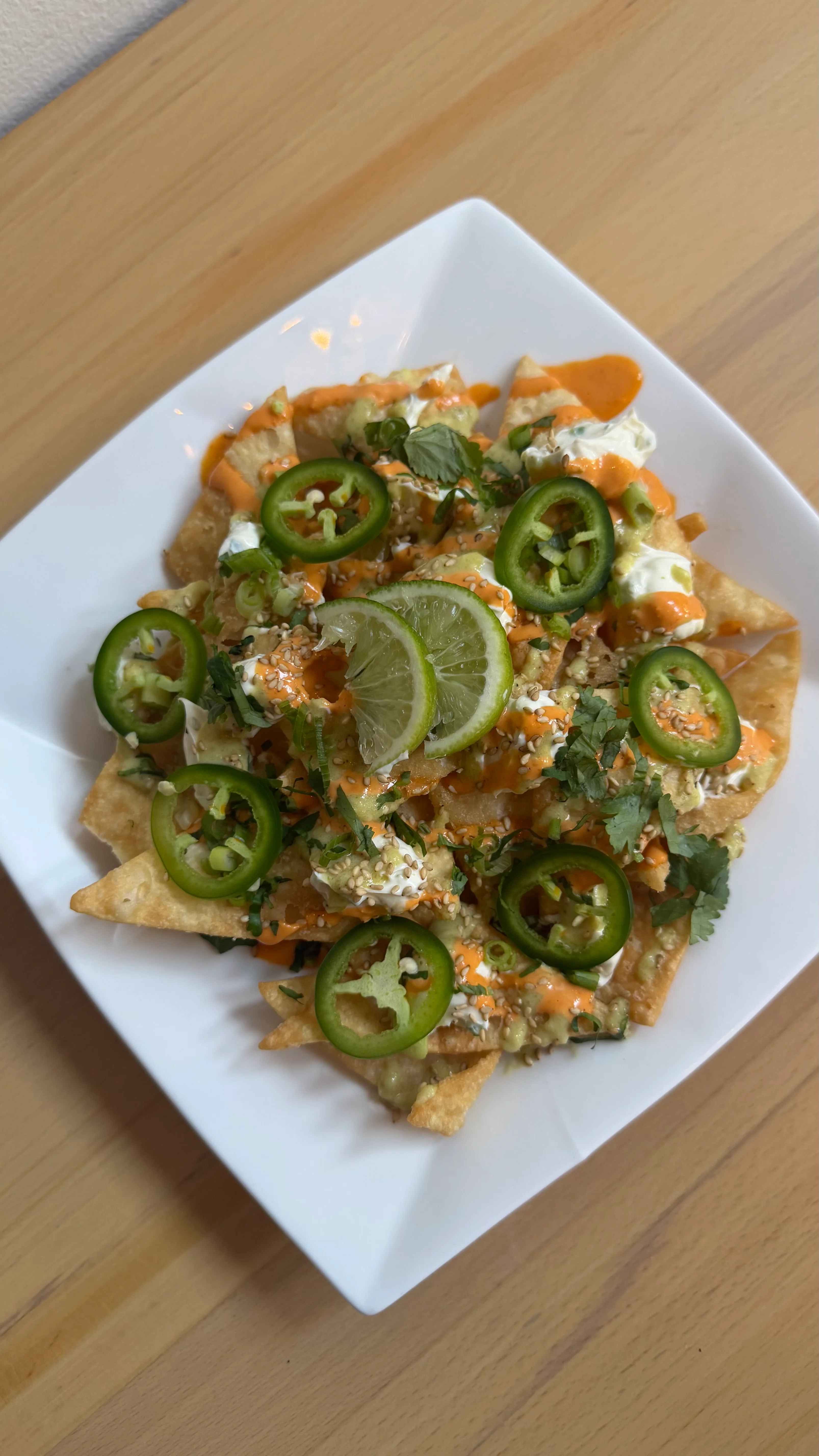 A plate of nachos topped with various ingredients, including jalapeños, sauces, lime, and fresh herbs. The nachos are arranged on a white square plate.