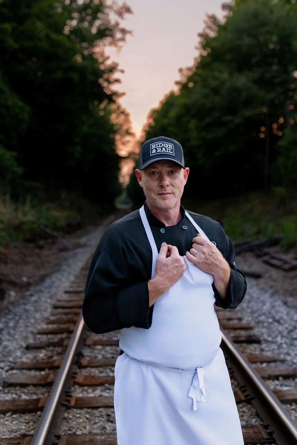 A chef standing confidently on a railway track, wearing a chef's coat and cap, surrounded by greenery.