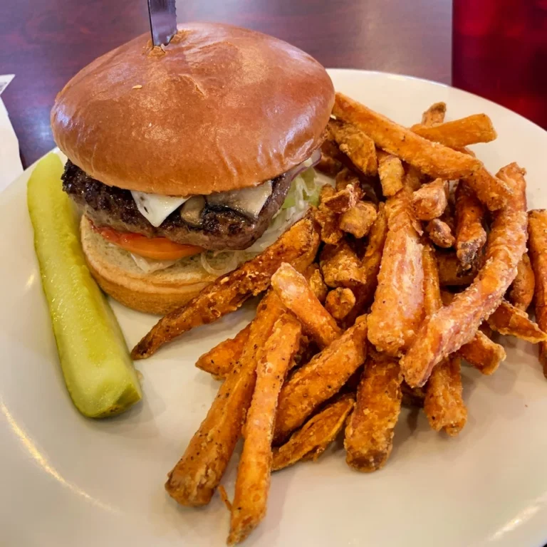 A juicy burger with lettuce, tomato, cheese, and mushrooms on a bun, accompanied by a generous portion of sweet potato fries and a pickle spear on a white plate.