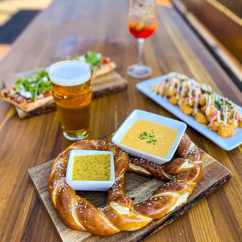 A large pretzel served with two types of dip, accompanied by a beer and other appetizers.