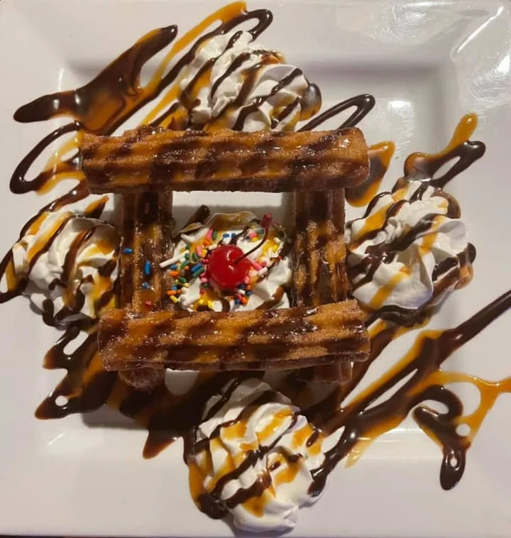 A dessert featuring churros arranged with ice cream, whipped cream, chocolate drizzle, and colorful sprinkles, topped with a cherry.