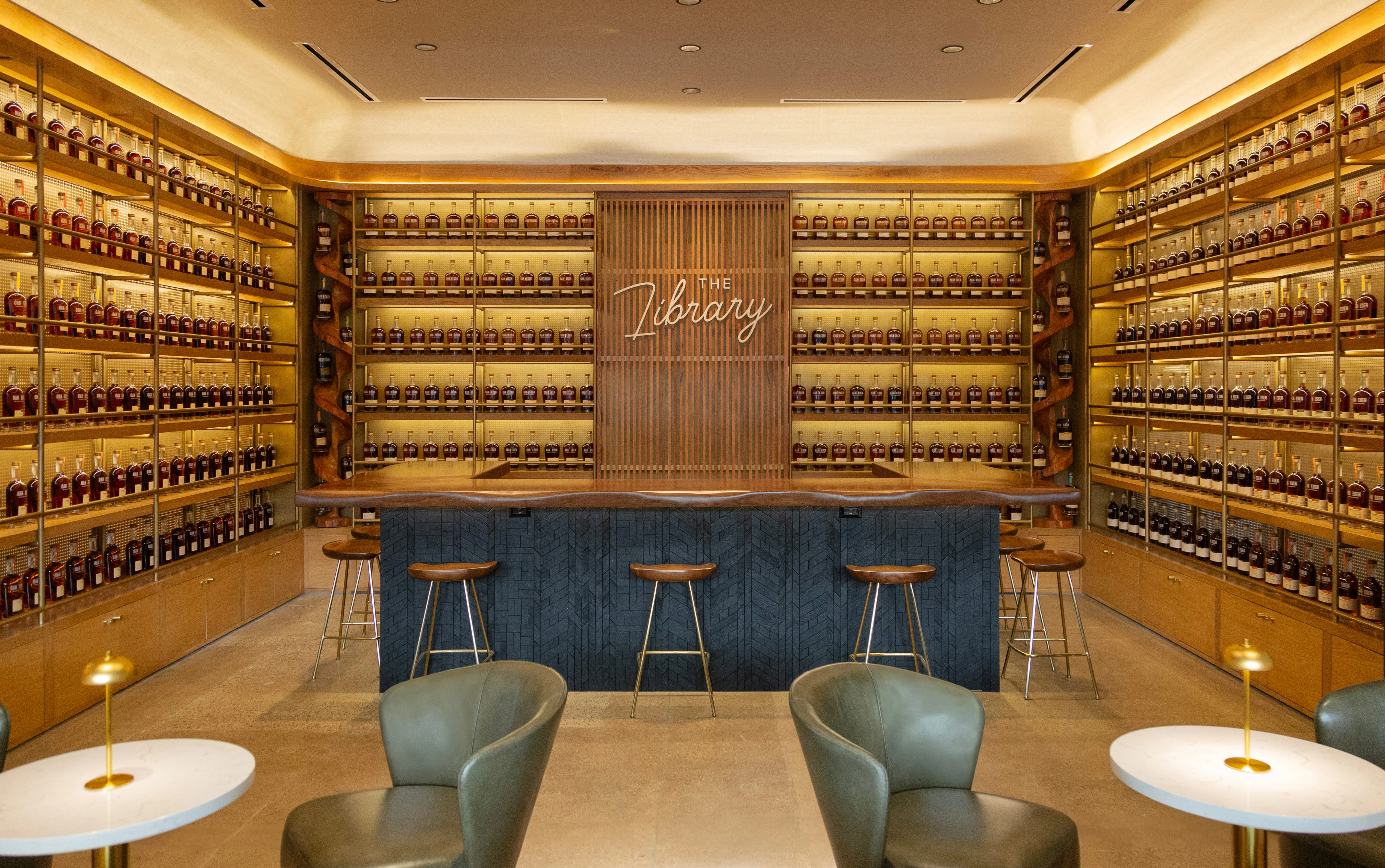 A stylish bar area featuring a long counter with wooden stools, surrounded by shelves filled with bottles. The walls are illuminated, highlighting the collection.