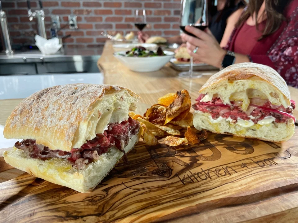 Two sandwiches on a wooden board, accompanied by potato chips. One sandwich is filled with beef and the other with assorted ingredients, with glasses of wine in the background.