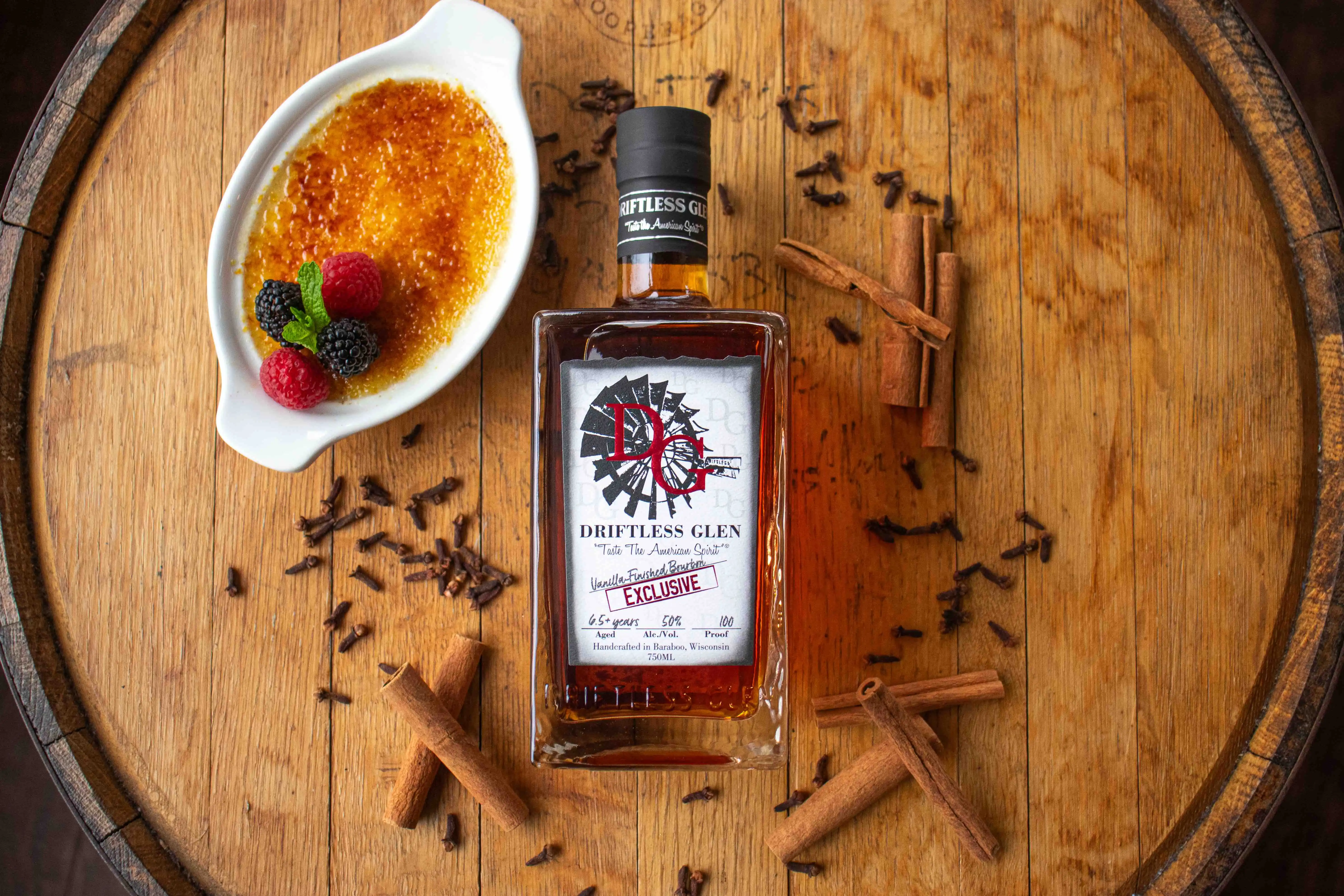 A bottle of Vanilla-Finished Bourbon sit on a whiskey barrel with cinnamon sticks and winter dessert around it.
