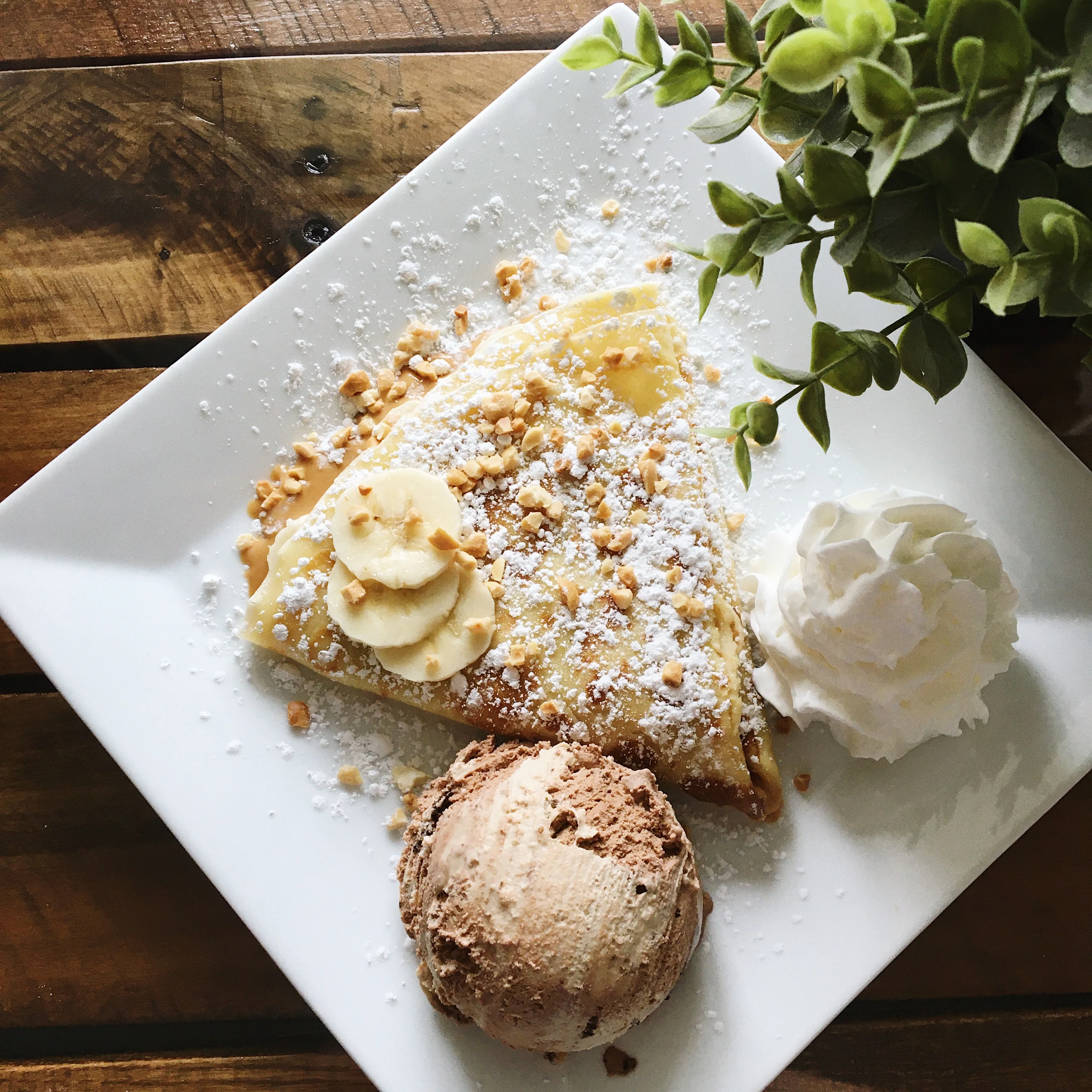 A delicious crepe topped with powdered sugar and crushed nuts, served with banana slices, whipped cream, and a scoop of chocolate ice cream on a square plate.
