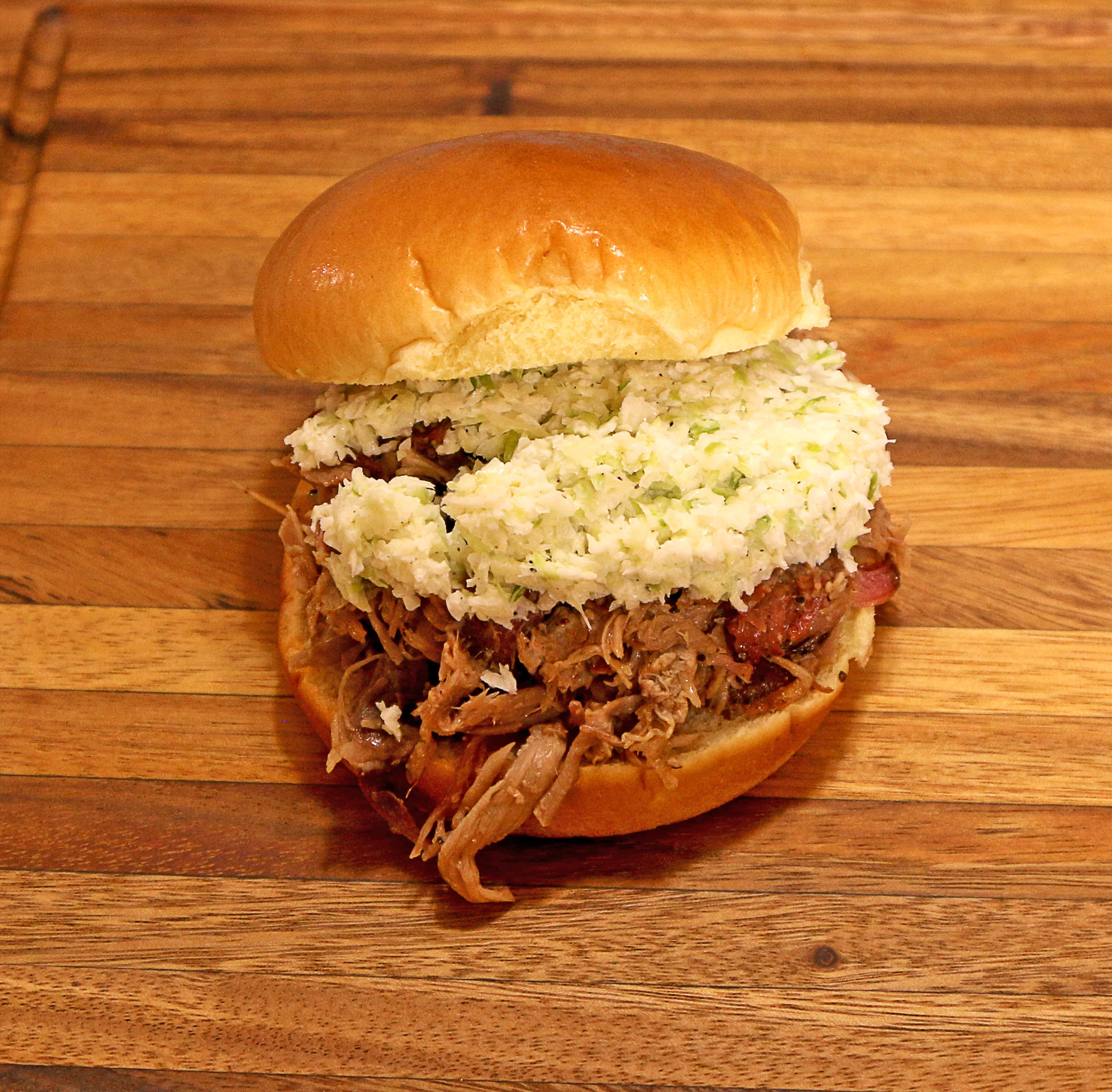 A wooden cutting board with a serving of pulled meat, baked beans, cheesy macaroni, and a dinner roll arranged neatly on it.