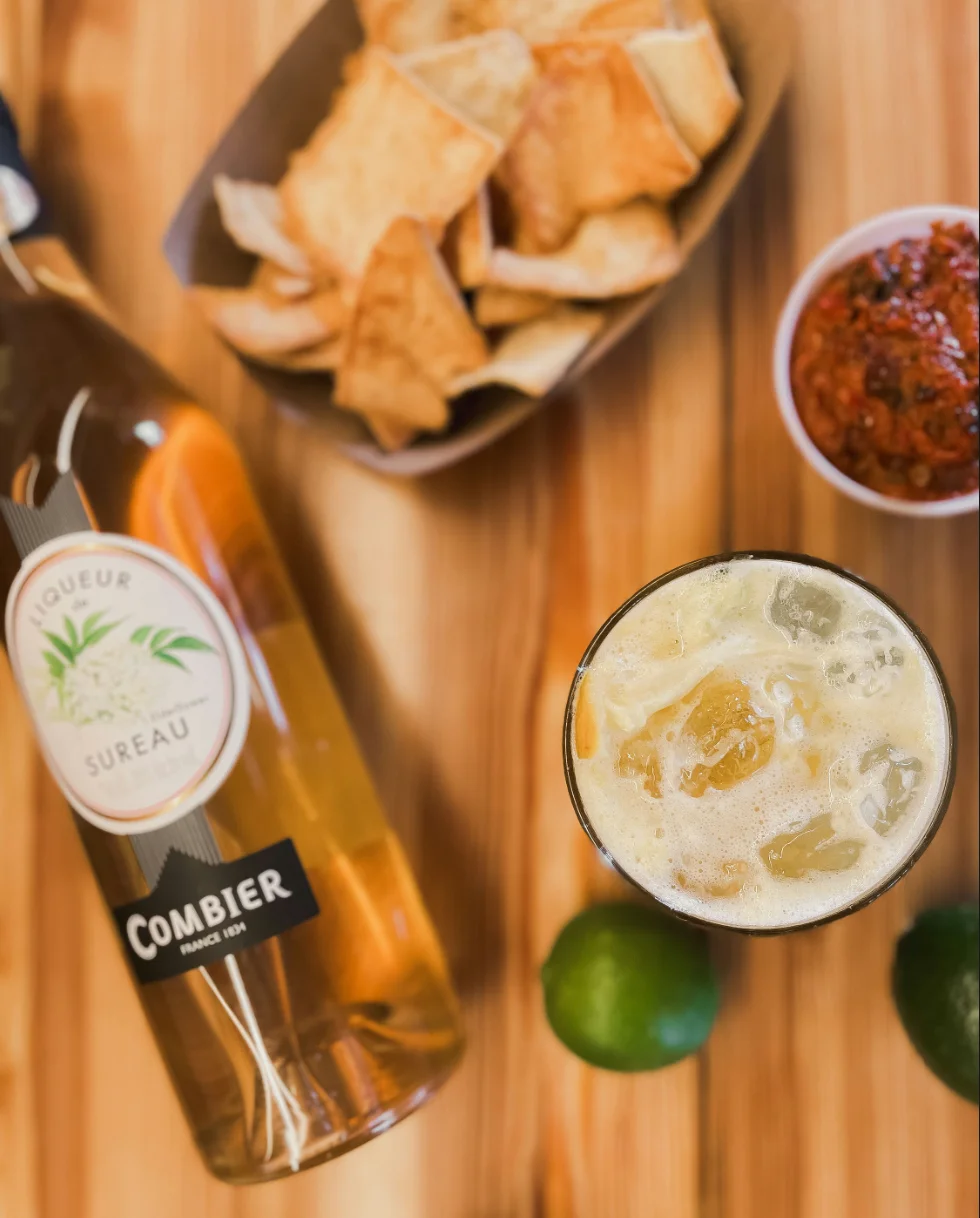 A refreshing drink with ice, lime, and a bottle of liqueur beside crispy snacks.