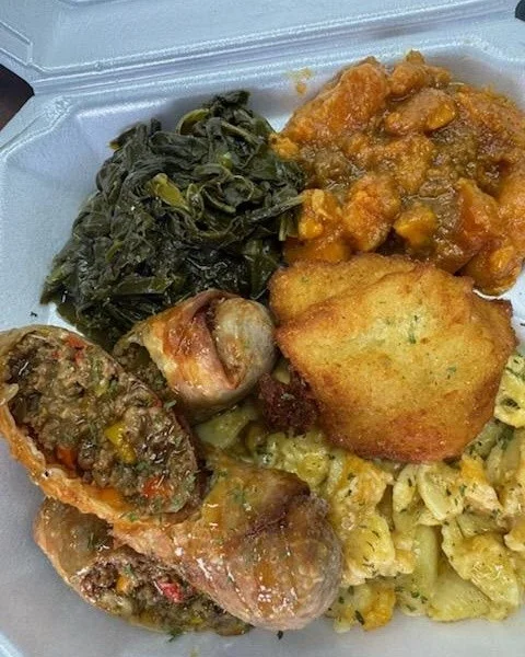 A takeout container filled with a variety of soul food items including greens, sweet potatoes, and meat rolls.