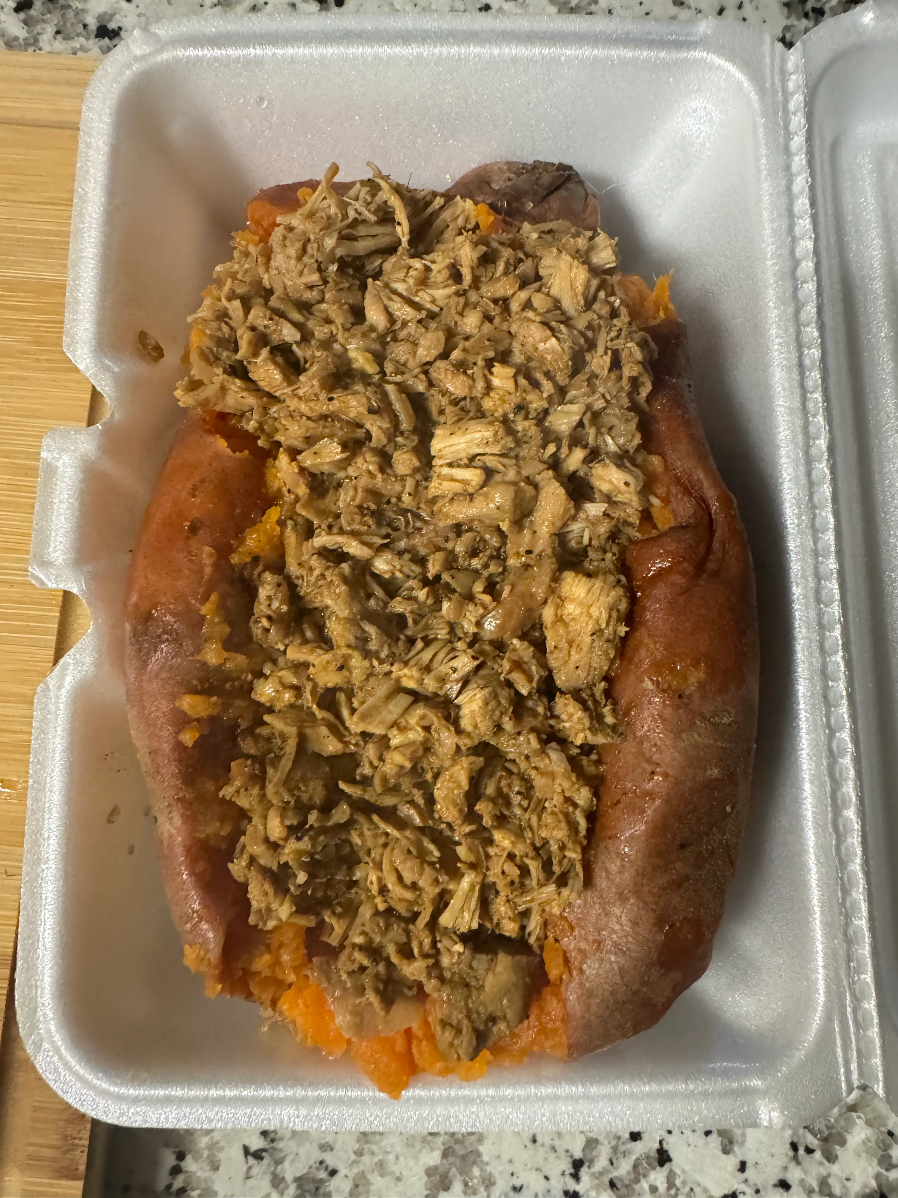 A baked sweet potato topped with shredded meat, served in a styrofoam container.