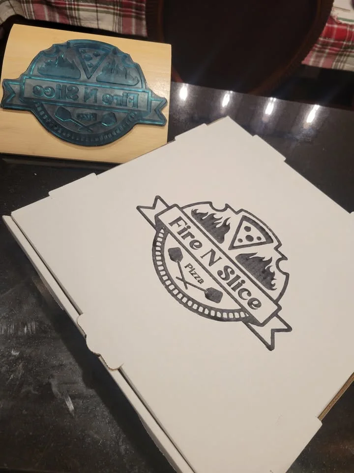 A pizza box featuring a logo with flames and a pizza motif. The box is white and sports a stylized design.