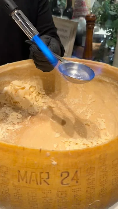 A large wheel of cheese with a blowtorch being used to melt the surface, revealing the inner texture.