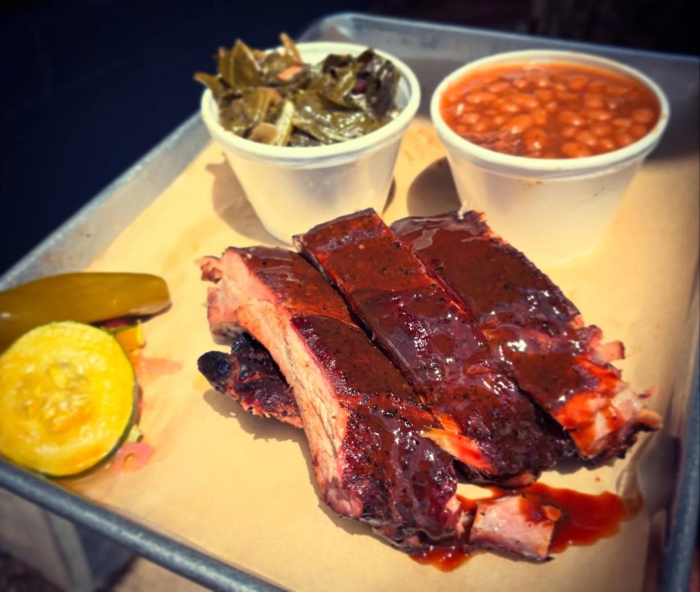 A plate featuring smoked ribs glazed with BBQ sauce, accompanied by pickles, collard greens, and baked beans in separate cups.