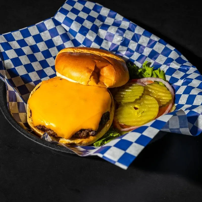 A loaded cheeseburger with a thick slice of melted cheese on a toasted bun, accompanied by lettuce, pickles, and tomato, in a checkered basket.