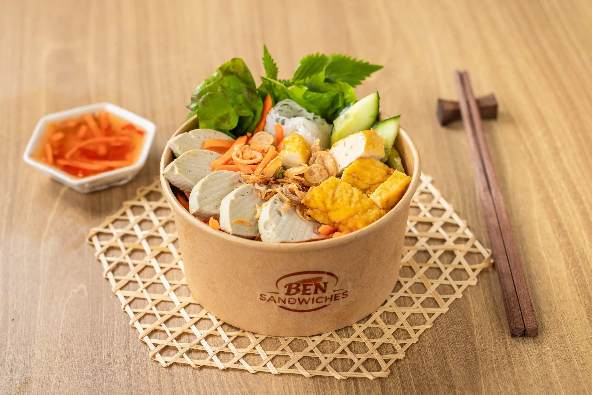 A bowl filled with a diverse assortment of ingredients, including noodles, vegetables, and fried toppings. Served with a side of dipping sauce.