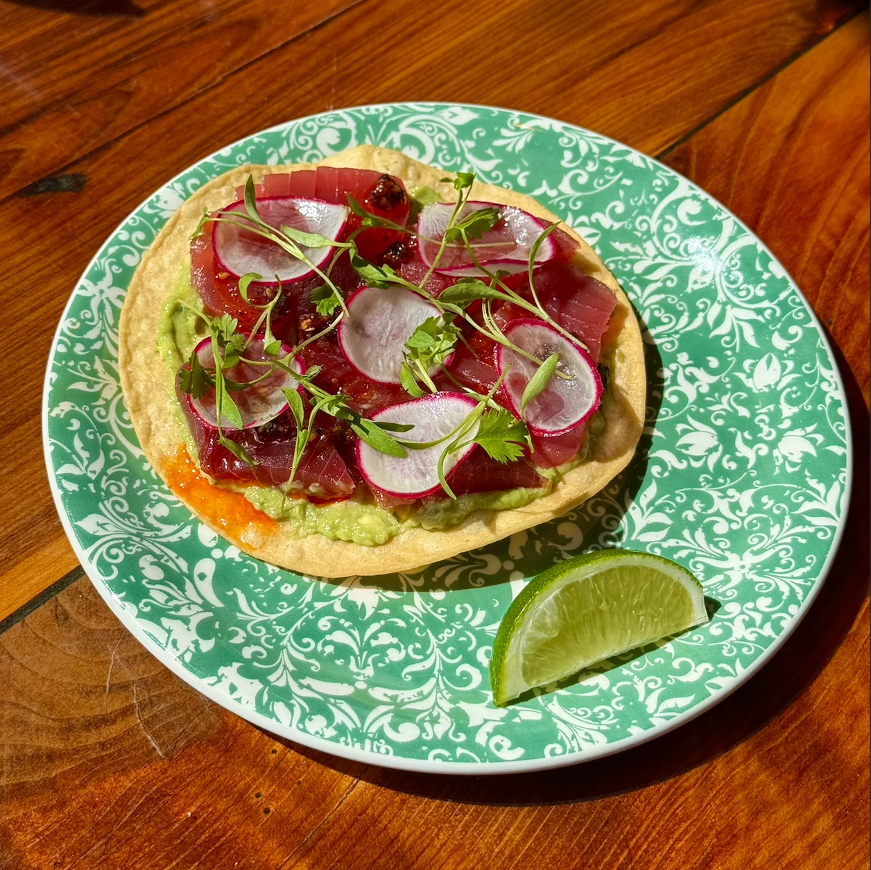 Tostada topped with fresh tuna, avocado, radishes, and lime on a decorative plate.