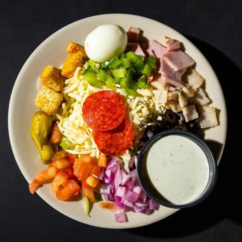 A colorful salad featuring mixed ingredients such as cheese, vegetables, meat, and a boiled egg, served with a dressing on the side.