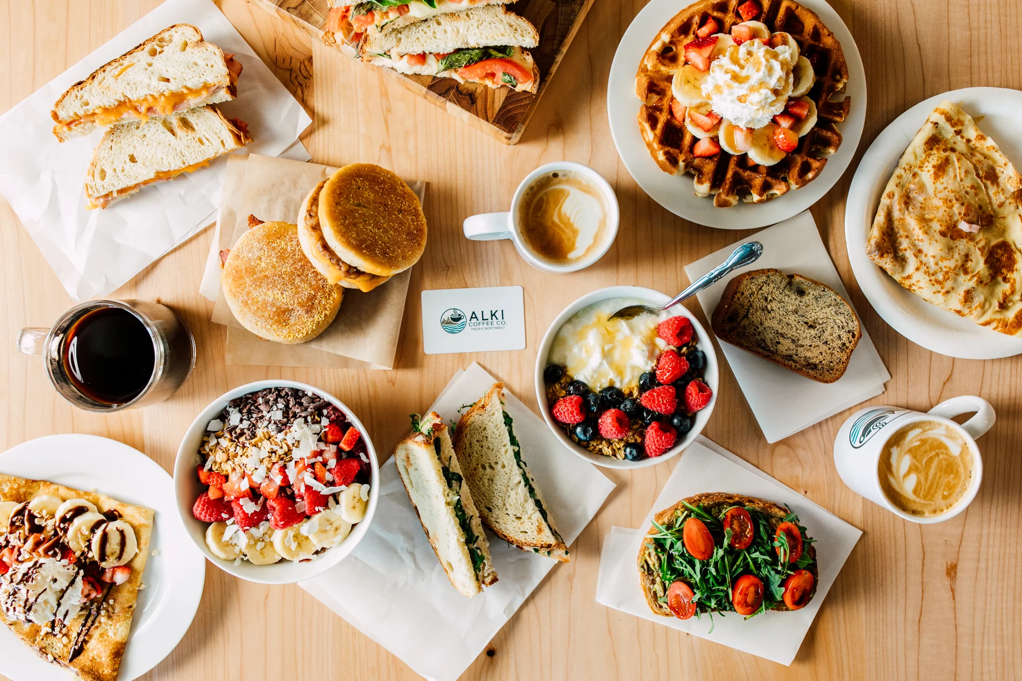 ssorted breakfast and coffee offerings at Alki Coffee Co., including espresso drinks, breakfast sandwiches, waffles, avocado toast, and fresh café favorites displayed on a wooden table.