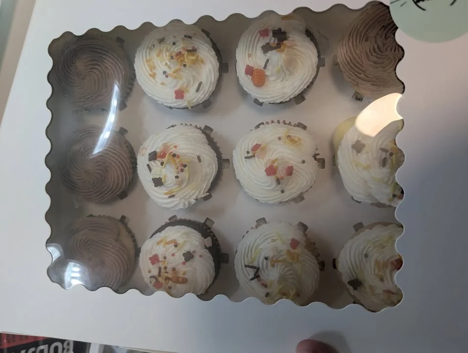 A box containing twelve decorated cupcakes with various frosting styles, some topped with colorful sprinkles and candy pieces.