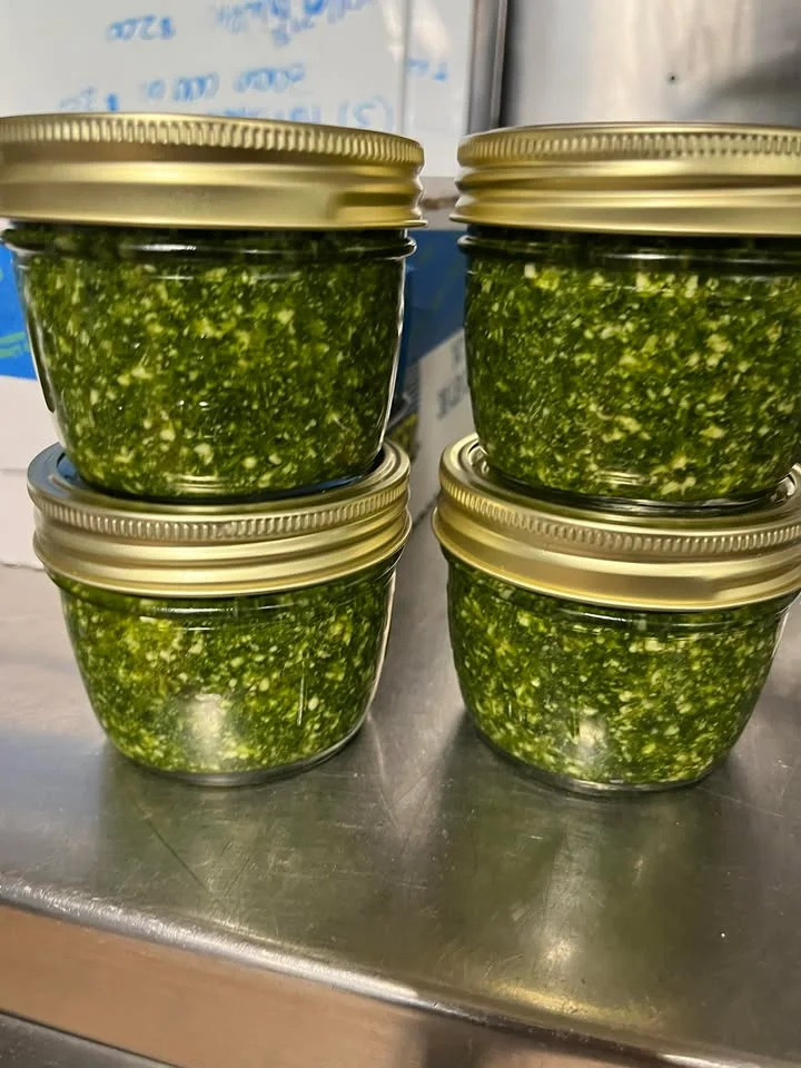 Four glass jars filled with a vibrant green pesto sauce, tightly sealed with golden lids, arranged on a metallic surface.