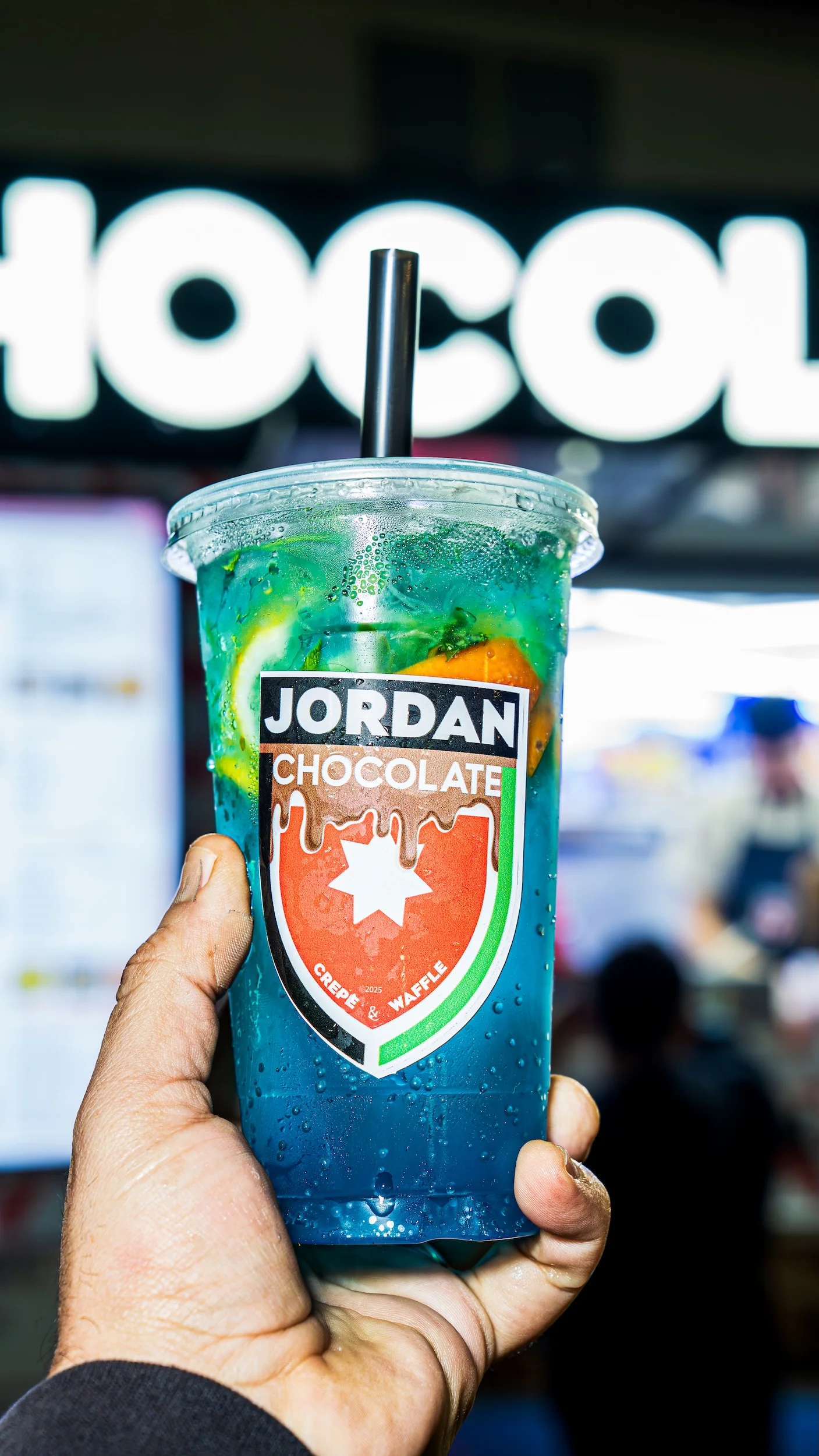 A vivid blue drink served in a clear cup, adorned with slices of lemon and mint. The drink features a prominent logo.