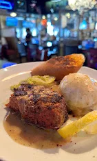 A serving of seasoned fish with lemon, accompanied by mashed potatoes, a fried item, and pickles.
