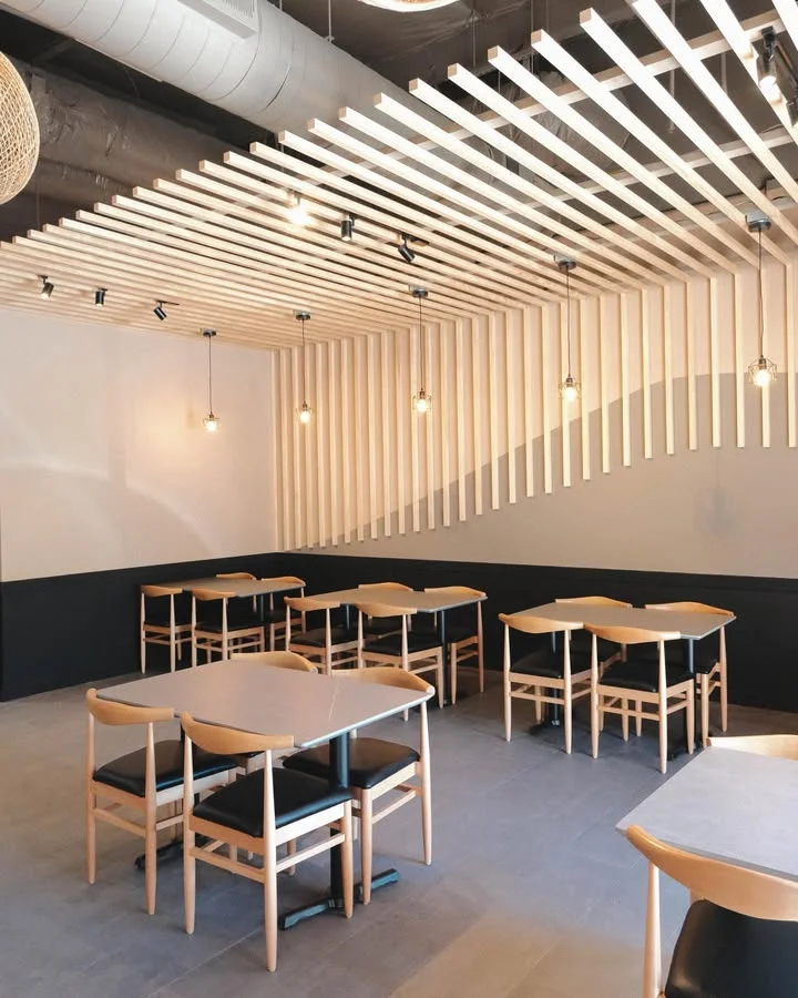 An empty dining space featuring wooden tables and chairs with a modern decor style. A unique ceiling design adds character to the space.