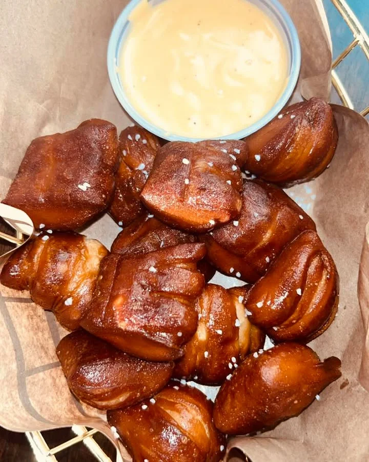 A basket filled with soft pretzel bites sprinkled with salt, accompanied by a small bowl of cheese dip.