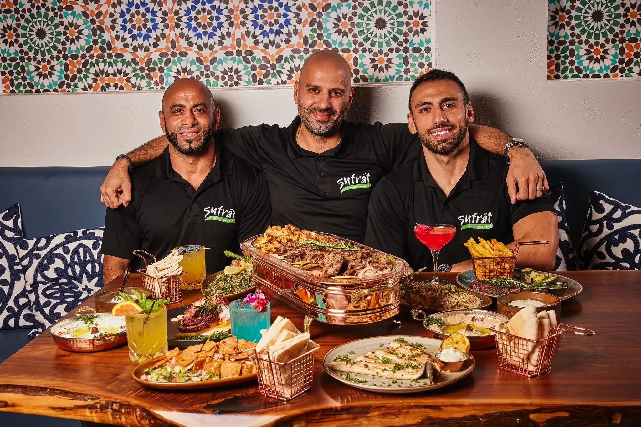 Three smiling men, the founders of sufrat in black branded sufrat shirts sit at a table filled with colorful Middle Eastern dishes and drinks, set against a backdrop of intricate tile patterns.