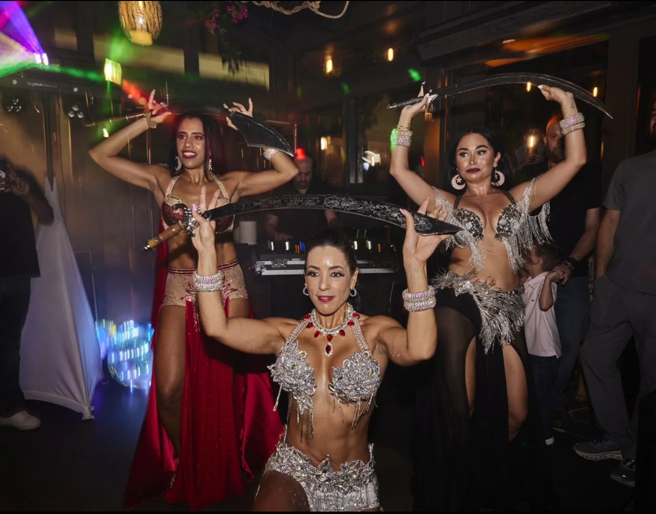 Three women in ornate belly dance costumes gracefully balance swords on their heads during a lively indoor performance, with colorful lights and an audience in the background.