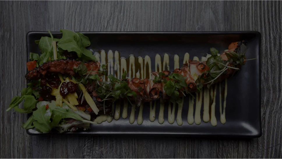 A top-down view of a grilled octopus tentacle garnished with microgreens, arranged over thin drizzles of a creamy sauce on a long black rectangular plate, accompanied by a fresh side salad with greens and mixed toppings.