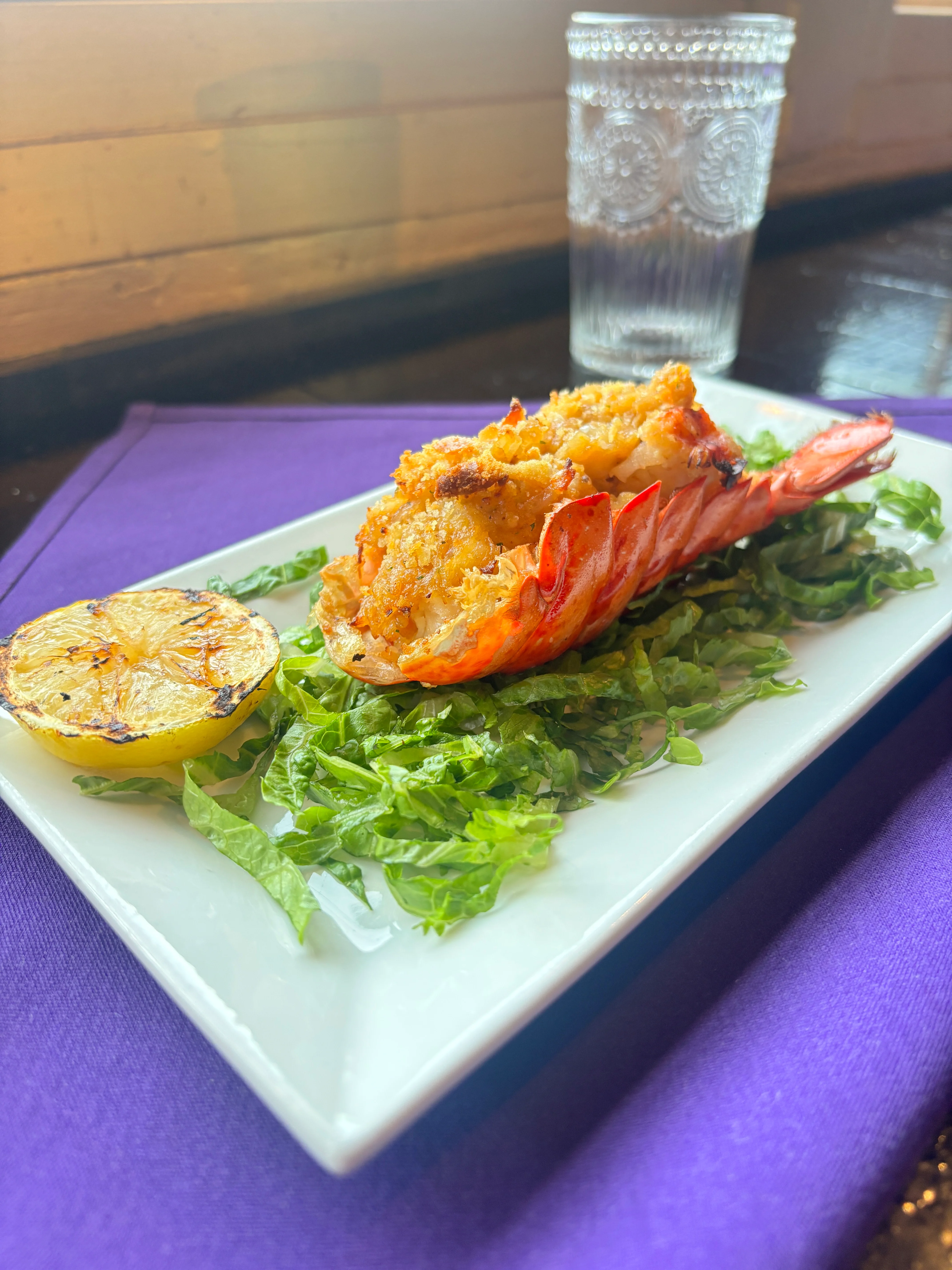 A gourmet plate featuring a large lobster tail, stuffed and baked, served on a bed of fresh greens with a grilled lemon slice.