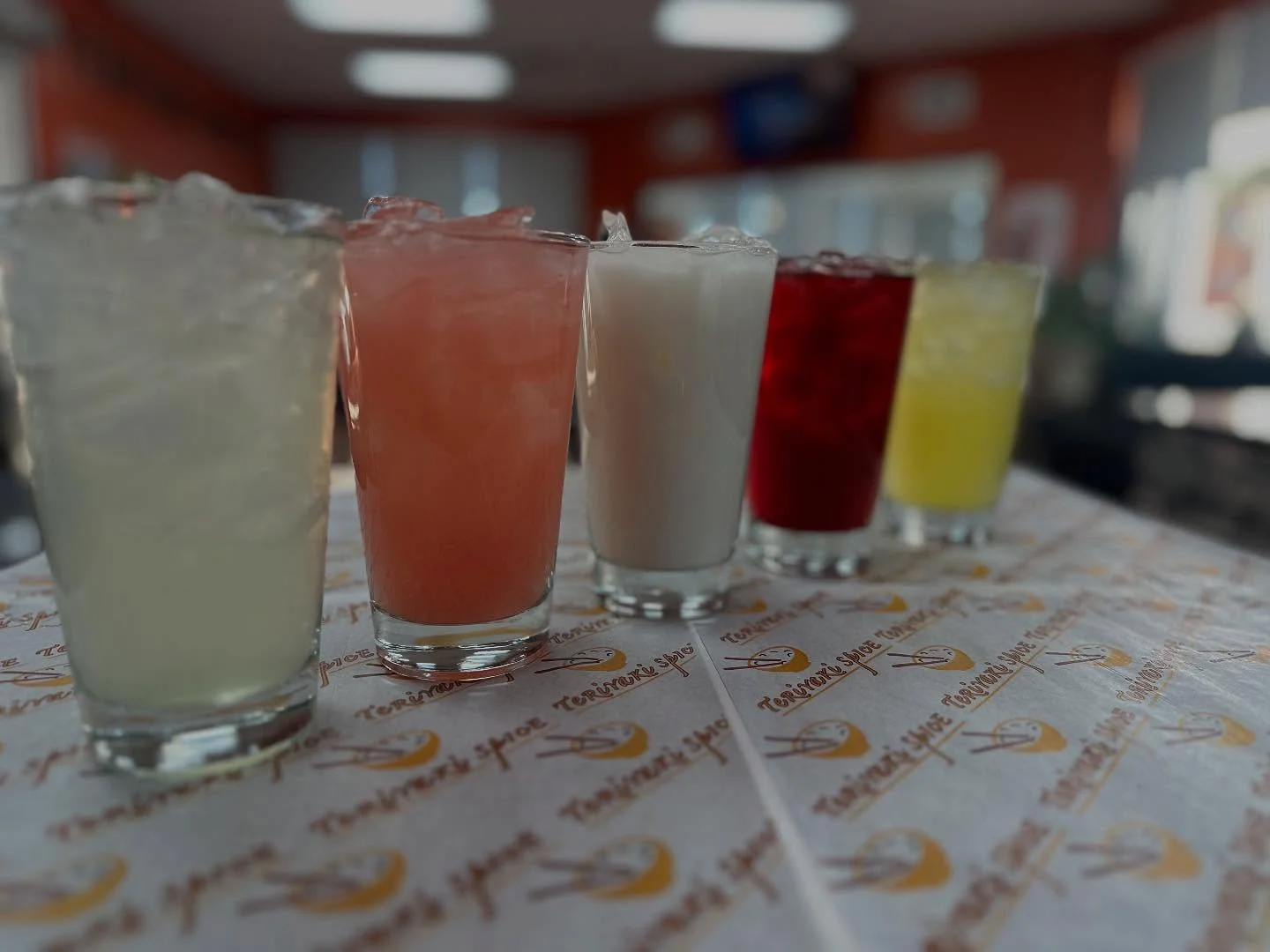 A row of colorful drinks with ice, including lemon, pink, white, red, and yellow variations, placed on a patterned tablecloth.