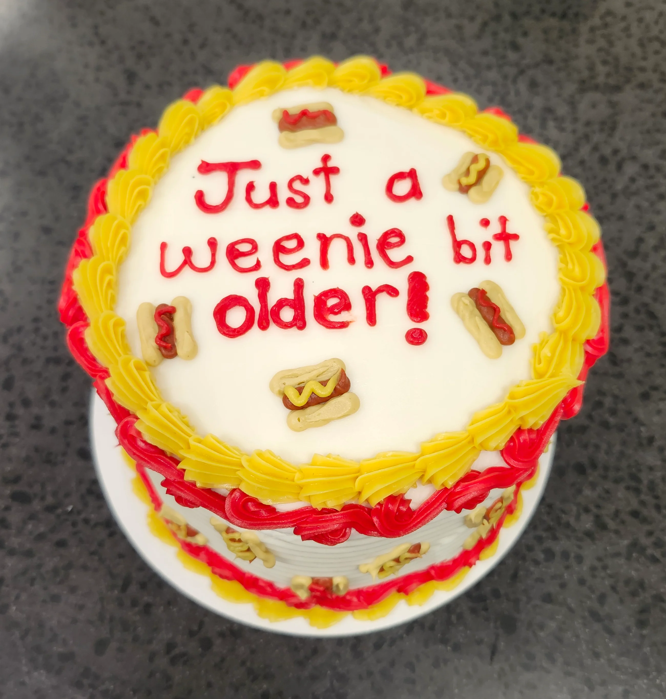 Small round cake decorated in yellow and red with tiny hot dogs that says "Just a weenie bit older!"