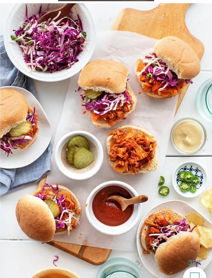 A collection of pulled pork sandwiches on a table, accompanied by purple slaw, pickles, and various sauces.
