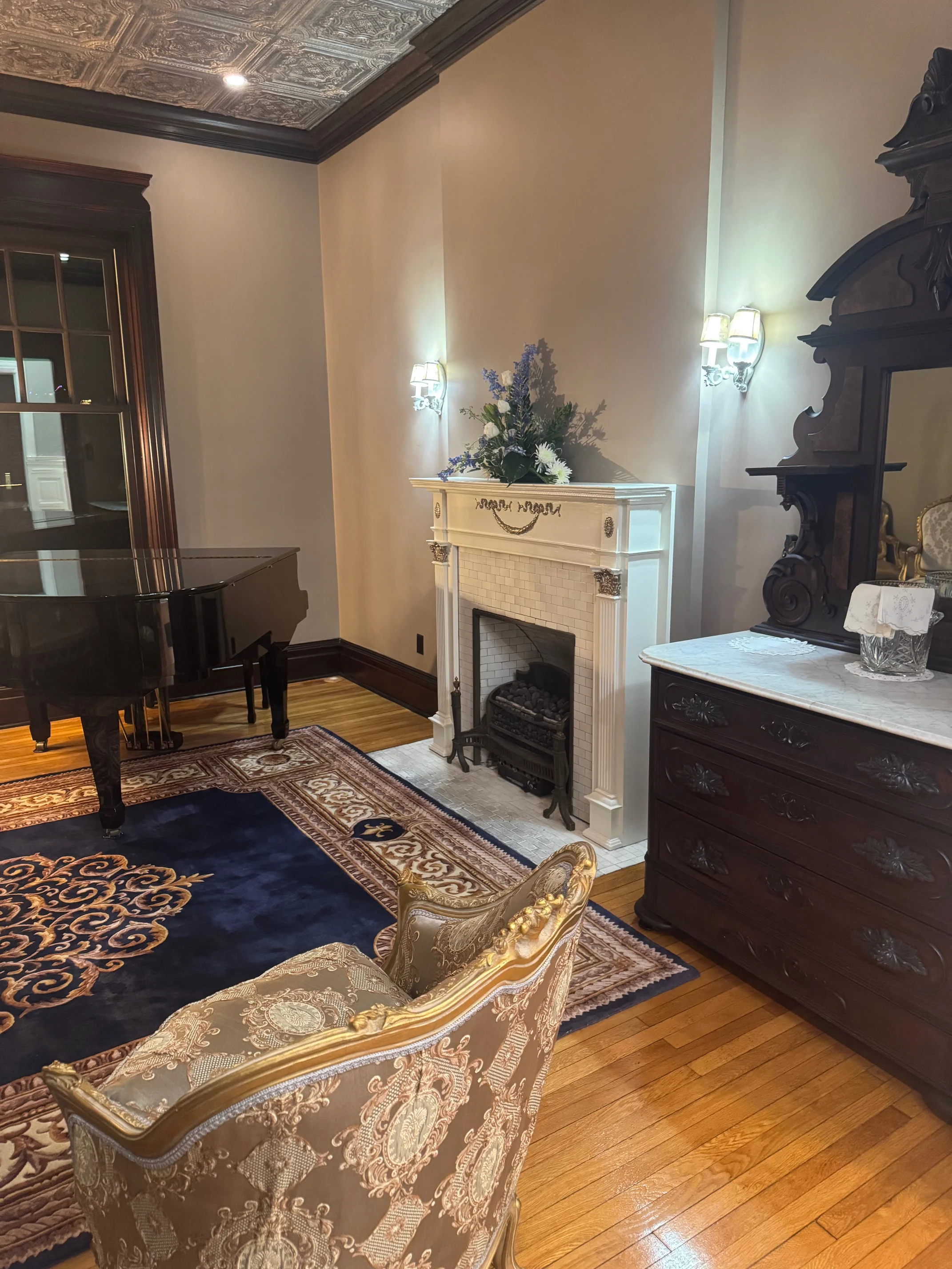 A cozy corner of a room featuring a grand piano, elegant armchair, and a decorative fireplace adorned with flowers. The woodwork and ceiling design add to the charm.