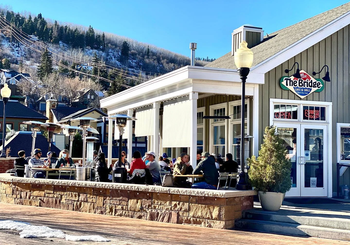 An outdoor dining area at a restaurant with patrons enjoying their meals while seated at tables. The setting features patio heaters and is surrounded by snow-capped mountains.