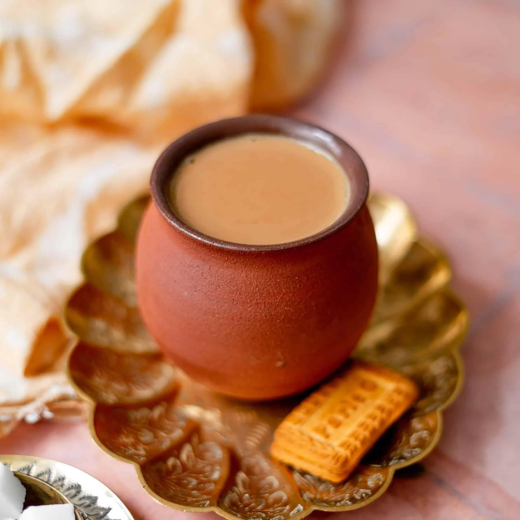 A clay cup filled with tea, placed on a decorative plate with a small biscuit beside it.