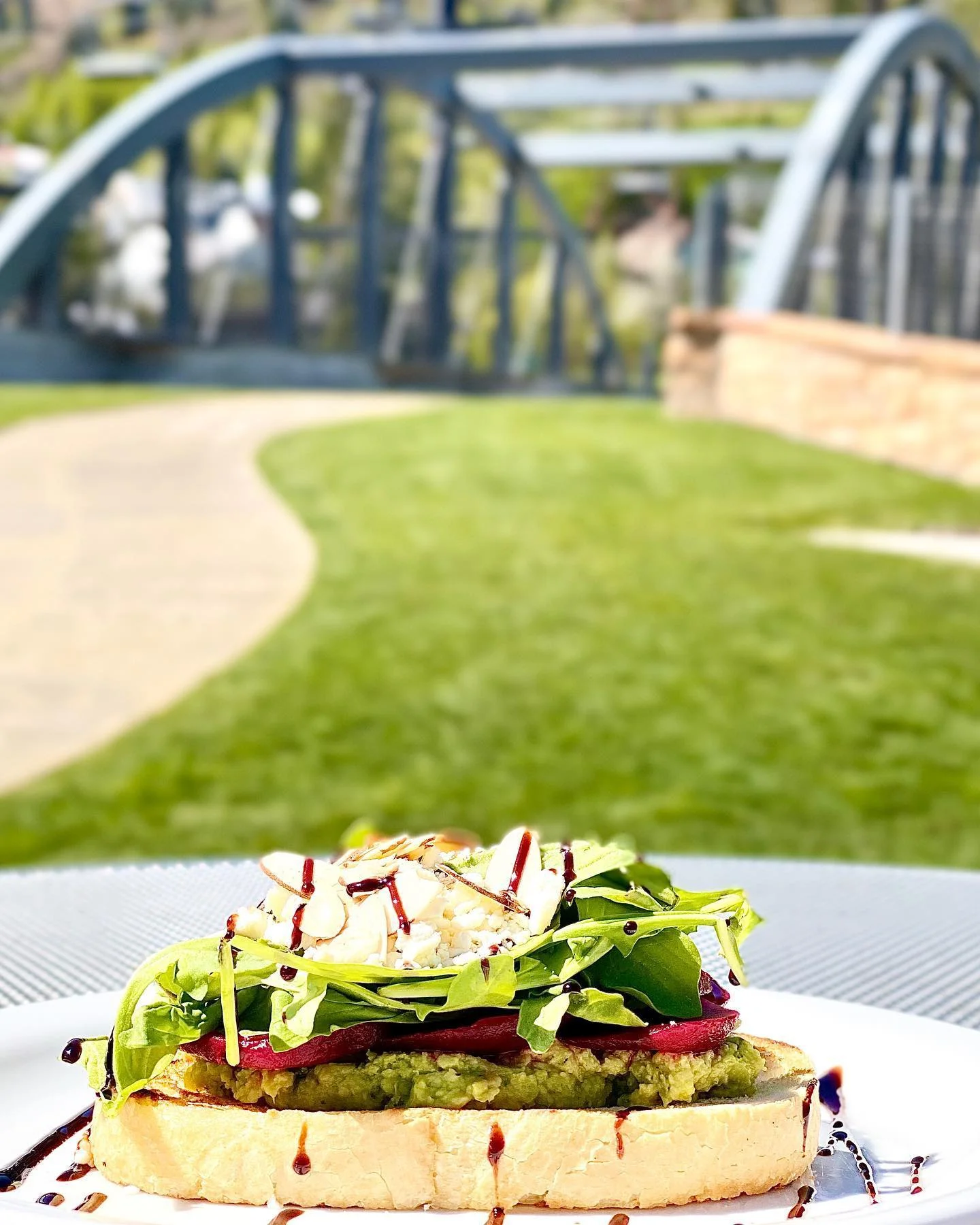 A slice of toasted bread topped with guacamole, spinach, beets, and sprinkled with almonds, set against a backdrop of a bridge and green grass.