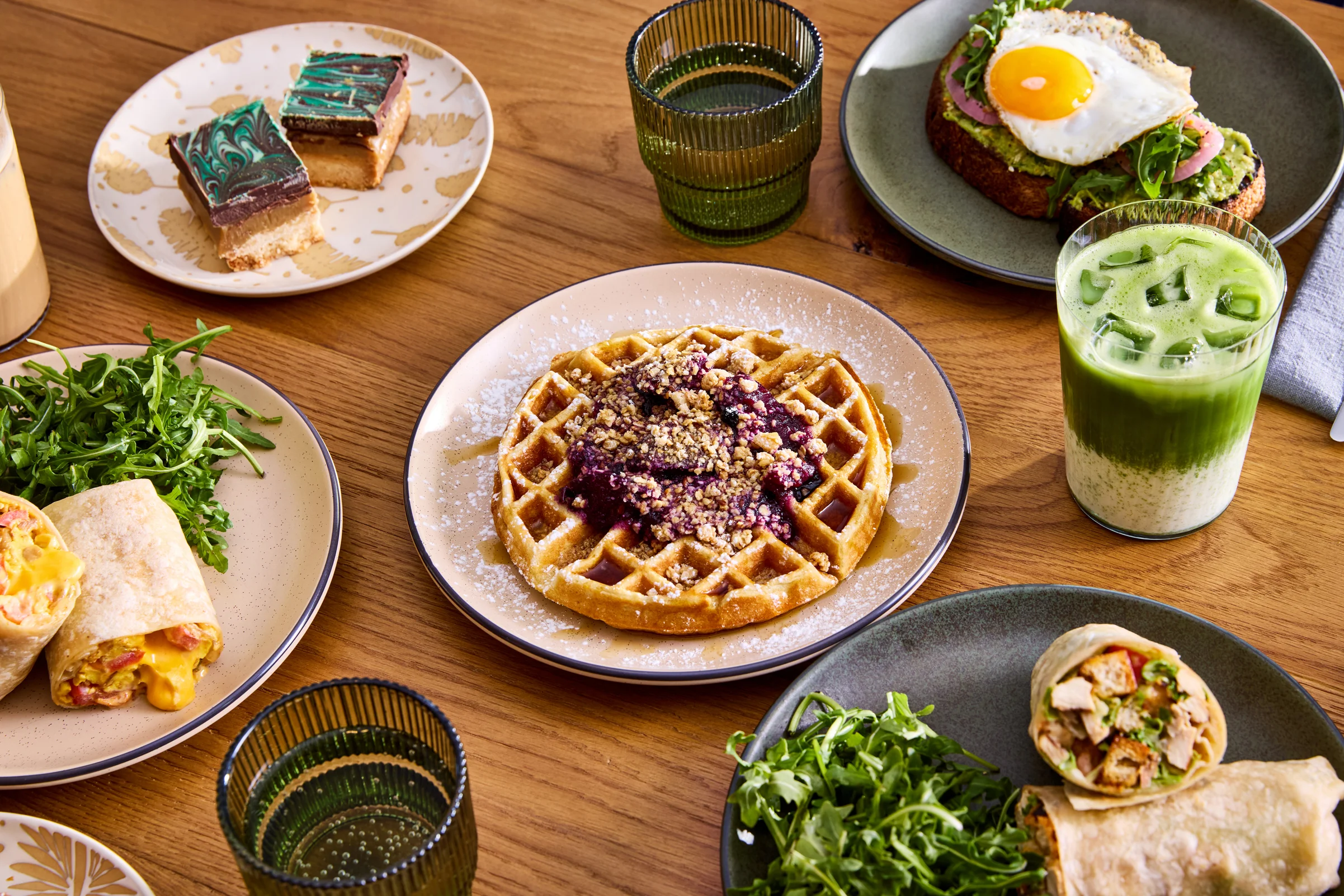 A delicious breakfast spread featuring a waffle topped with berries and nuts, along with a wrap, greens, and a glass of green drink on a wooden table.