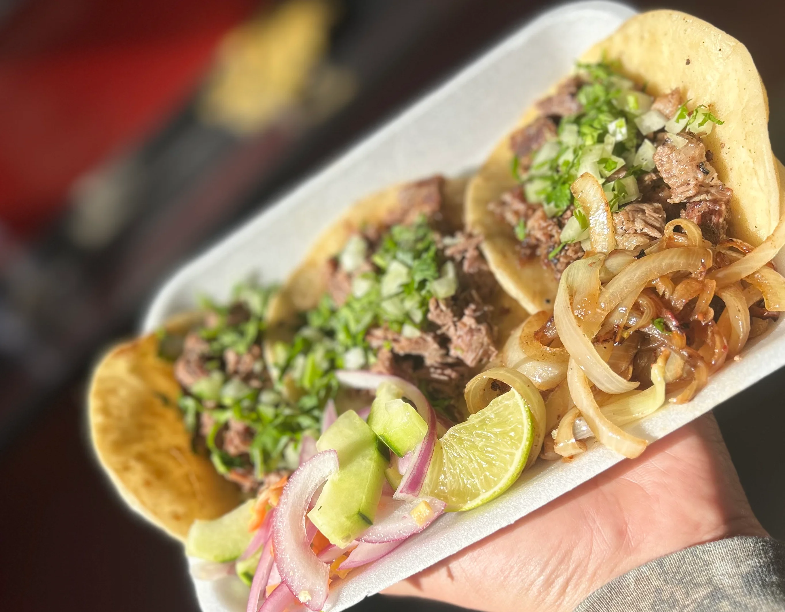 A large platter of assorted tacos, including toppings like onions and cilantro, served alongside fresh lime, cucumber, and radishes. The tacos are arranged neatly on a checkered tray.