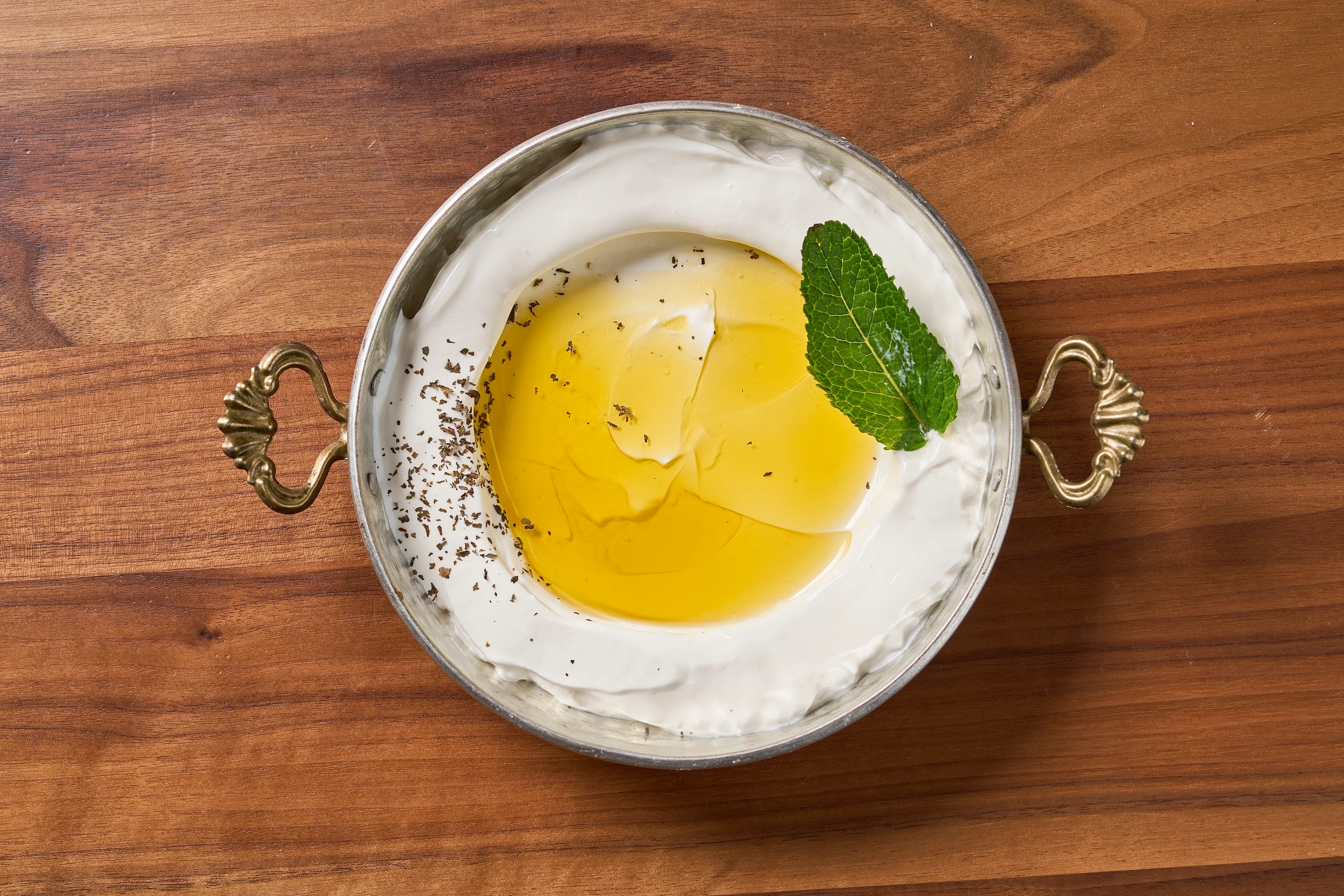 A metal dish with creamy yogurt topped with a drizzle of olive oil, a sprinkle of black pepper, and a mint leaf, on a wooden table. Elegant, fresh, rustic.