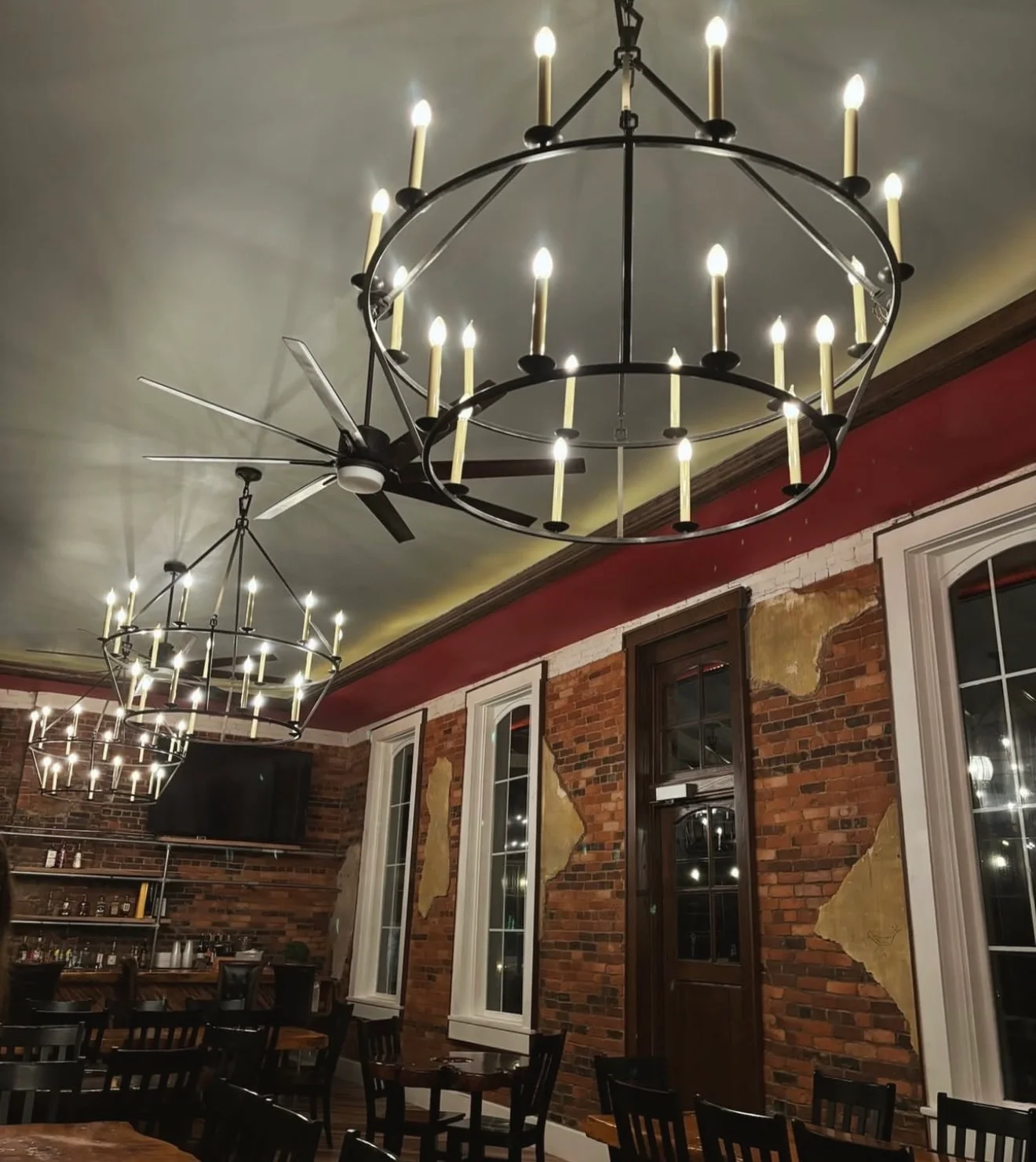 An interior view of a dining space featuring elegant chandeliers and exposed brick walls. The room is well-lit and has wooden furniture.