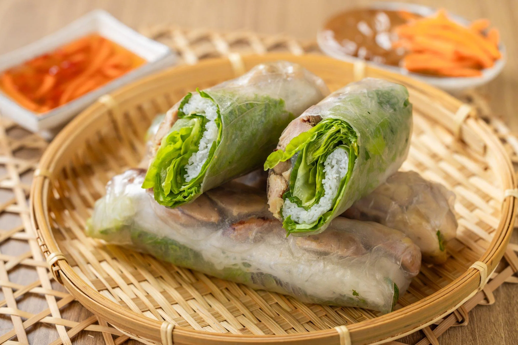 Rice paper wraps filled with lettuce and meat arranged in a bamboo basket. Condiments including sauce and pickled carrots are placed nearby.