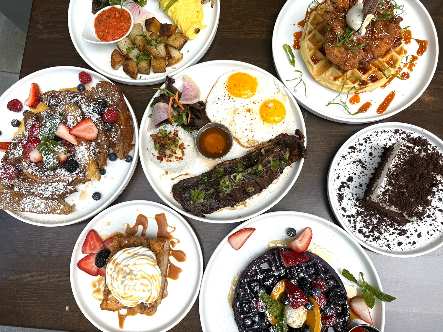 Overhead view of assorted brunch dishes at Sprout Cafรฉ in Palo Alto, featuring French toast, chicken and waffles, sunny-side eggs, ribs, fresh fruits, and a chocolate dessert
