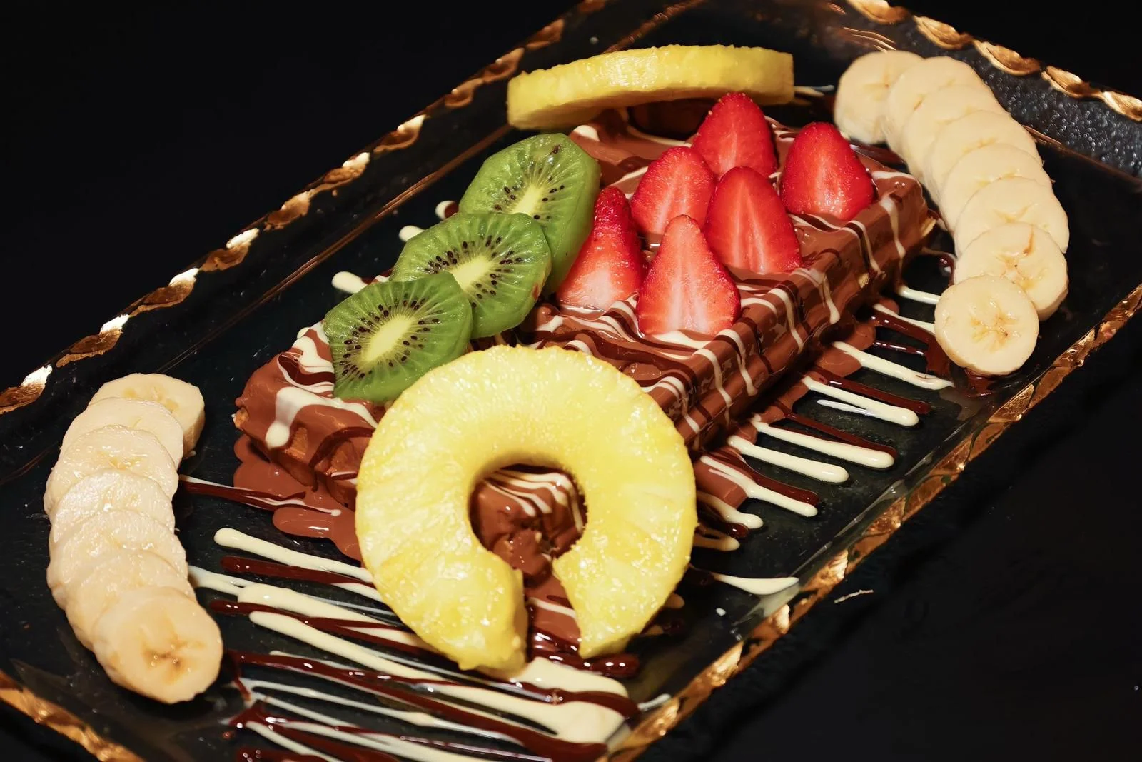 A decorative plate featuring a chocolate dessert topped with fresh fruits including strawberries, kiwi, banana, and pineapple, drizzled with chocolate sauce.
