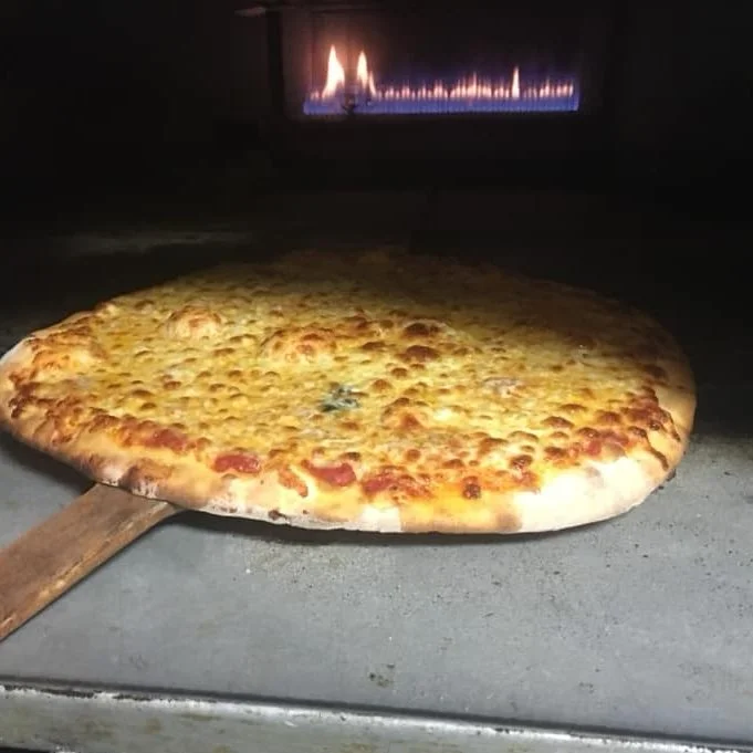 A freshly baked cheese pizza on a wooden pizza peel, with a background showing a wood-fired oven.